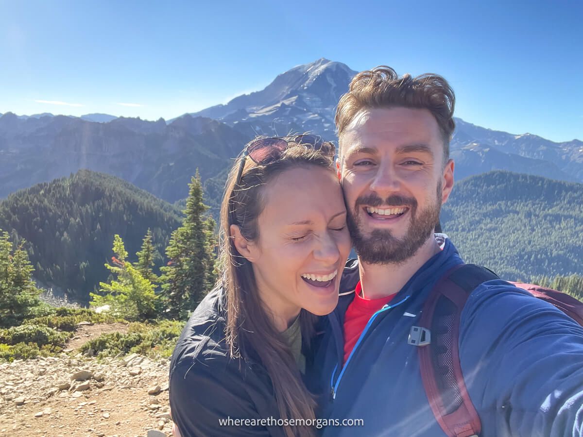 Photo of Mark and Kristen Morgan from Where Are Those Morgans laughing together at the summit of Tolmie Peak Trail with views of Mt Rainier behind on a sunny day with clear blue sky