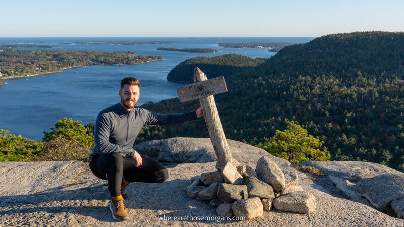Photo of Mark Morgan hiking in Acadia and crouching down to hold a wooden summit marker on a rocky surface with views over a lake behind 