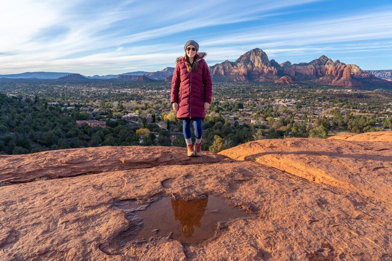 Photo of Kristen Morgan in winter coat on a table-like rock formation reflecting in a shallow puddle with far reaching views behind