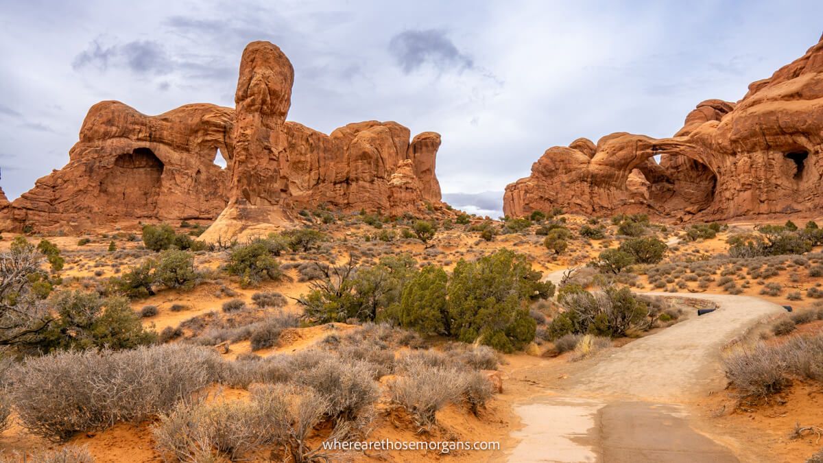Photo of a path leading through a red rocky desert to natural arch formations on a cloudy day in Utah