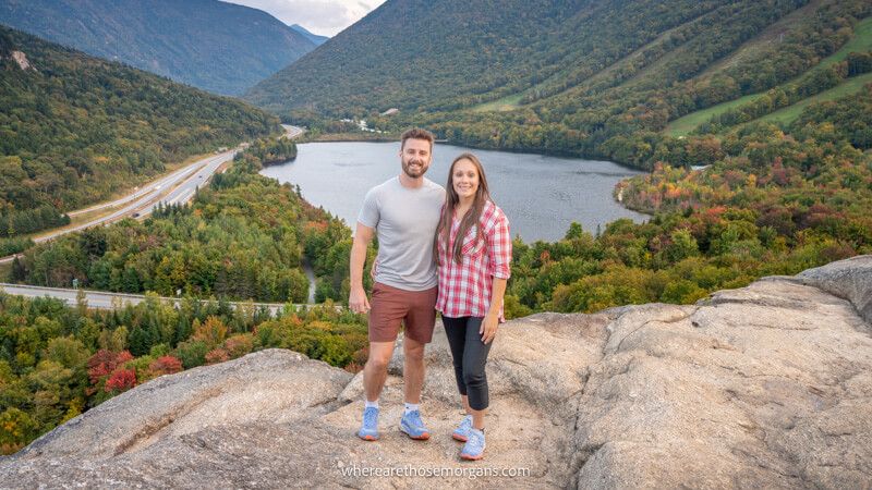 Photo of Mark and Kristen Morgan from Where Are Those Morgans standing together on a rocky summit with views over a lake and rolling hills covered in trees behind