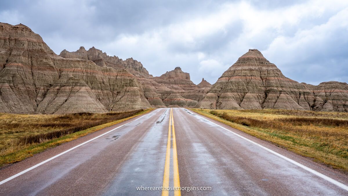An empty straight road leading into small granite mounds with red sediment layers under a cloudy sky