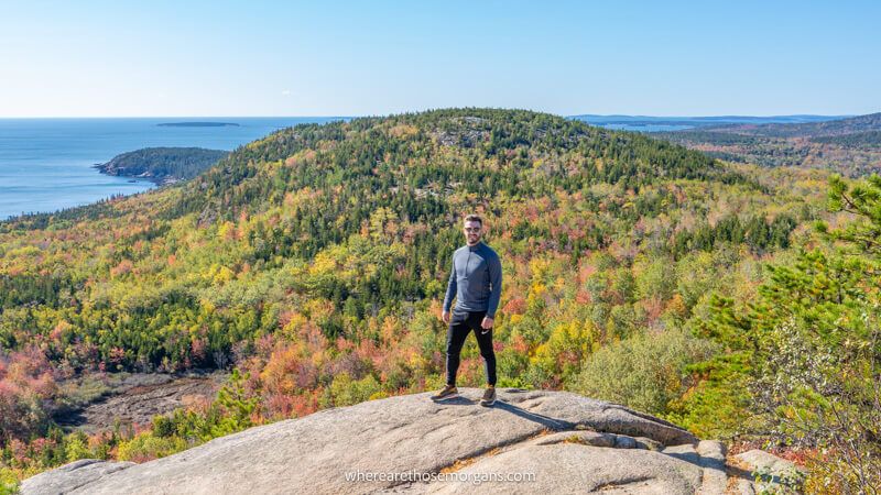 Photo of a hiker stood on a rocky summit plateau with views over rolling hills covered in colorful tree leaves and the ocean on a clear day in Maine