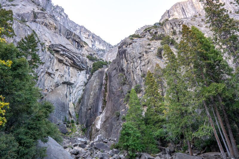 Photo of a dry waterfall eroded into a tall granite cliff face with evergreen trees at the base