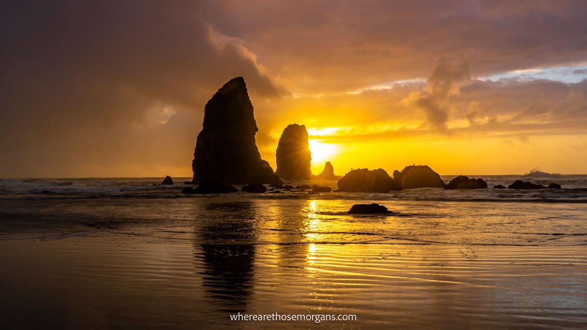 Rocky haystacks in shallow water in the ocean behind a beach covered in water and a sunset with thick clouds