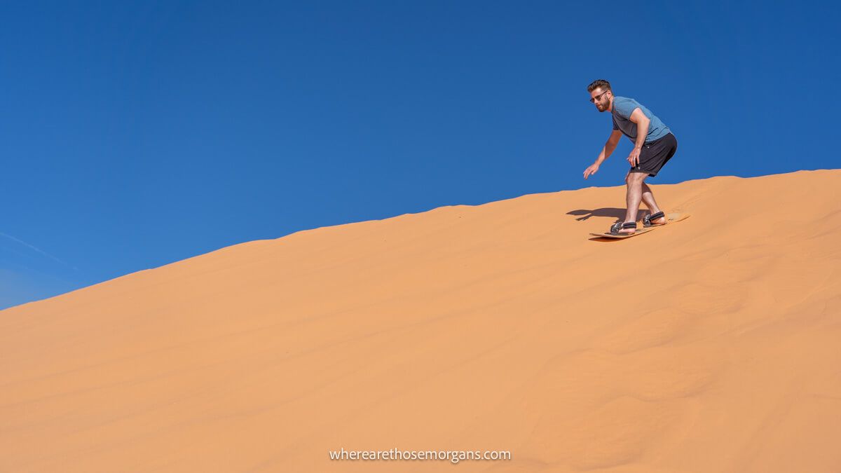Photo of a sand boarder in shorts and t-shirt sliding down a steep dune with a clear blue sky