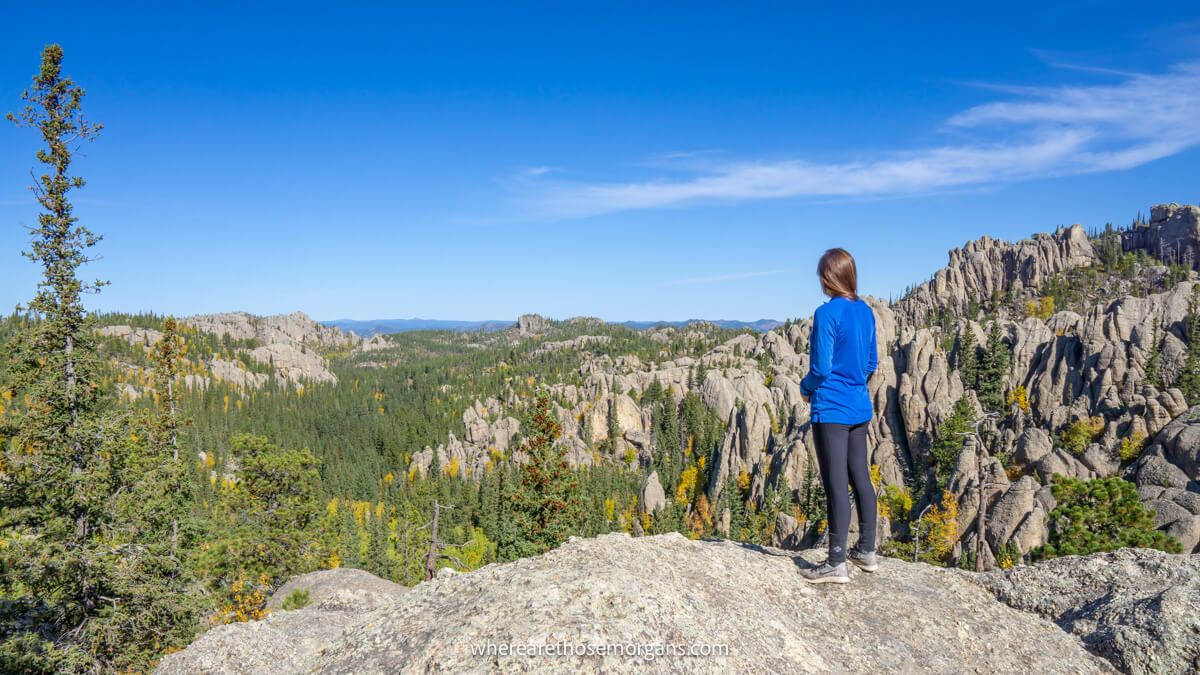 Photo of a hiker in a blue fleece stood on rocks looking out at views over a granite landscape covered in evergreen trees