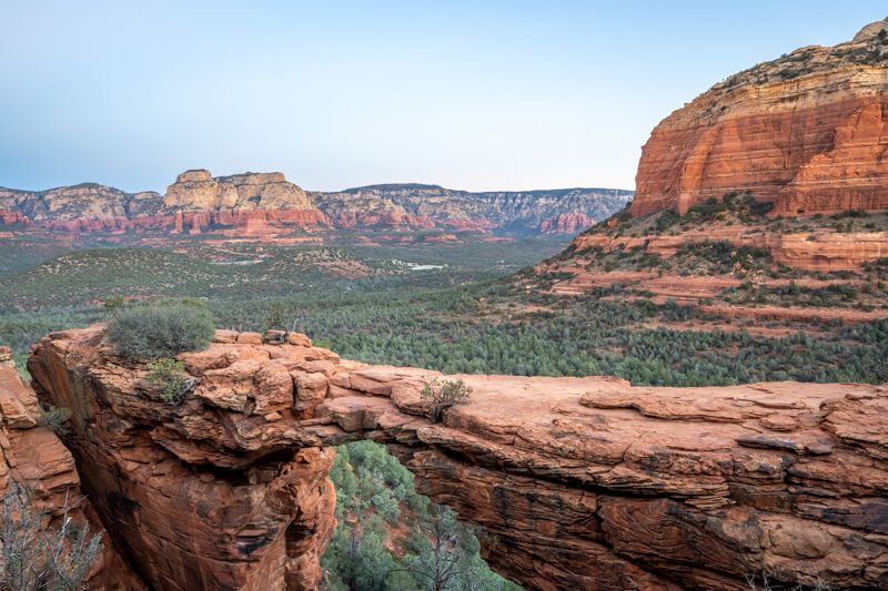 Photo of a bridge-shaped rock formation with far reaching views over a canyon floor filled with trees at dawn