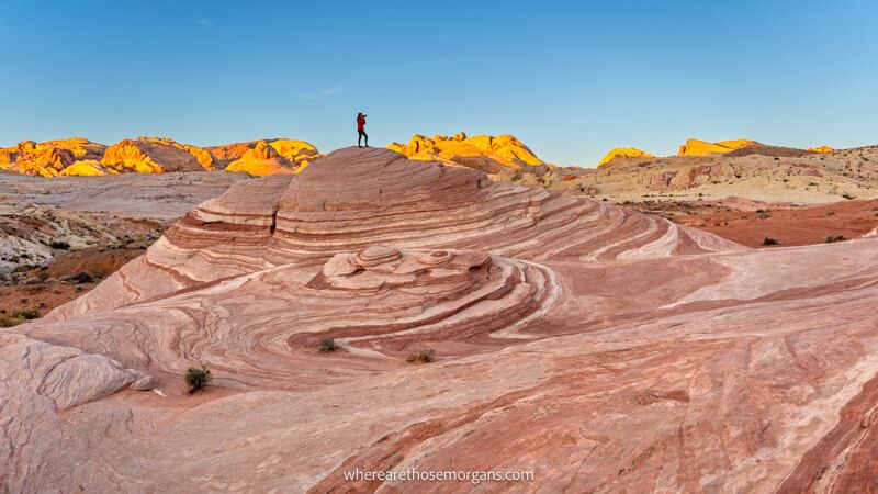 Photo of a hiker in the distance stood with a camera on top of a red and white domed rock formation with swirling patterns in the Valley of Fire in Nevada