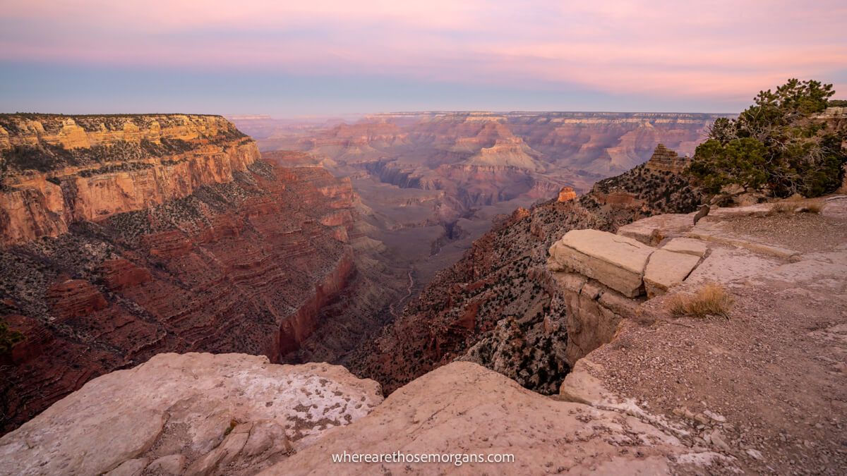 Photo of the top of a rocky rim with long drops down into a deep sandstone canyon with pink colors in the sky at dawn