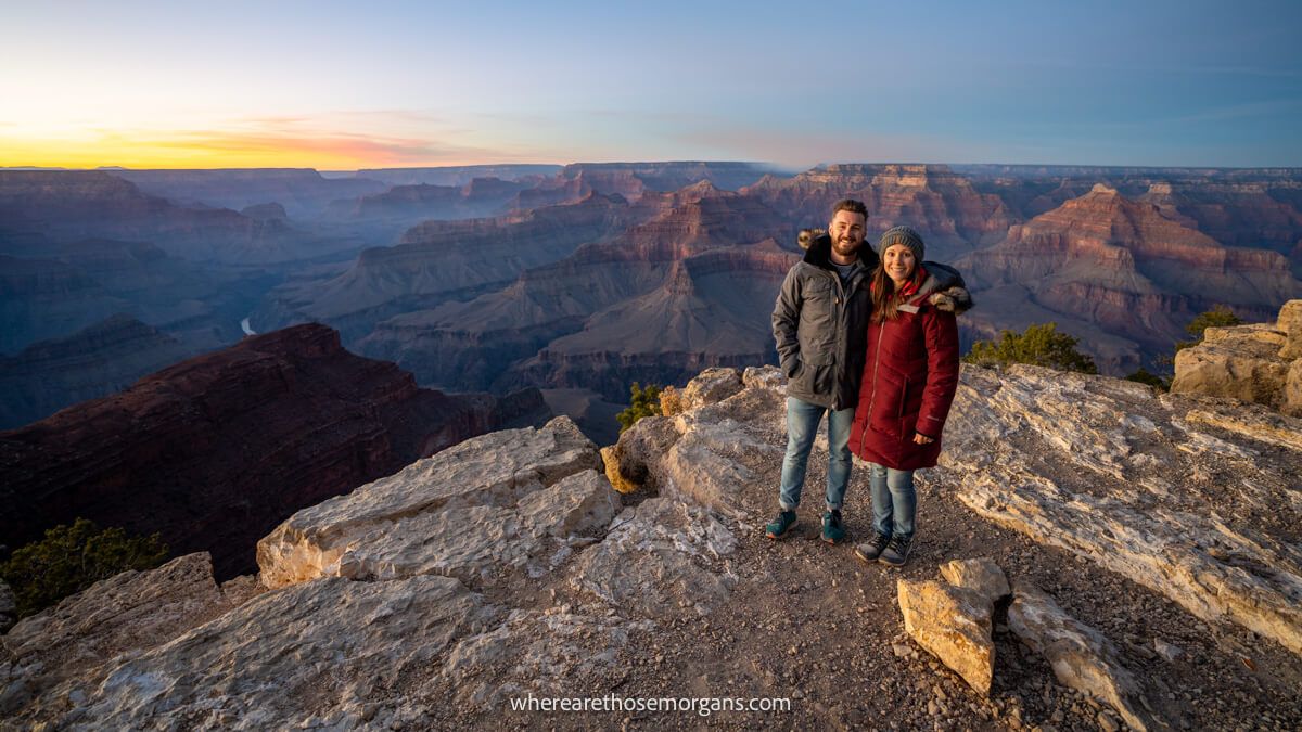 Couple standing together in winter coats on a rocky ledge with views into the Grand Canyon at sunset