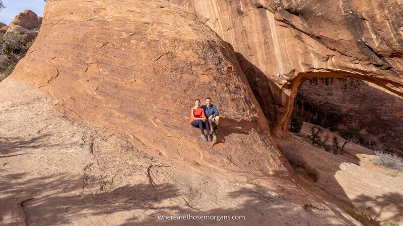 Photo of two hikers sat on a sloping sandstone rock next to a natural arch