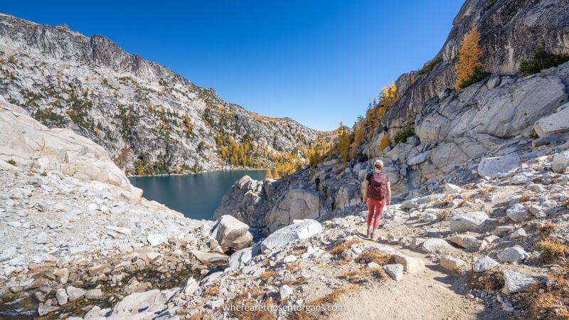 Photo of a person walking through a rocky landscape with a lake and golden larches under a clear blue sky