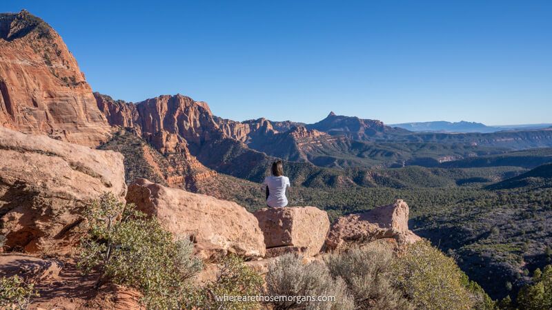 Photo of a hiker sat on a small red rock looking out at far reaching views of sandstone mountains and trees in a valley