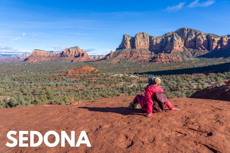 Photo of a hiker sat in winter clothes on a flat red rock mesa looking out at far reaching views of a canyon valley filled with trees under a blue sky and text reading Sedona