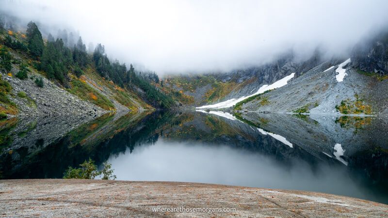Photo of a still lake reflecting rocks and snow with dense clouds above