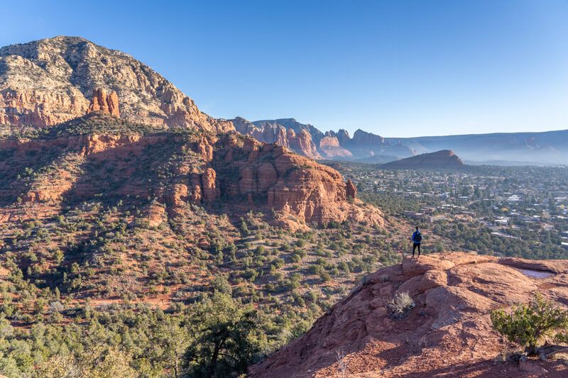 Photo of a hiker standing on a distant red rock flat shelf looking at views over deep valleys and rock formations under a blue sky in Sedona Arizona