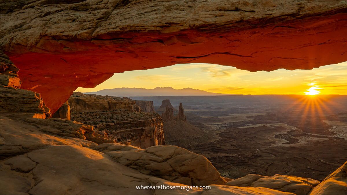 Natural arch rock formation shaped like an eye glowing red at sunrise with views of a vast flat canyon through the arch