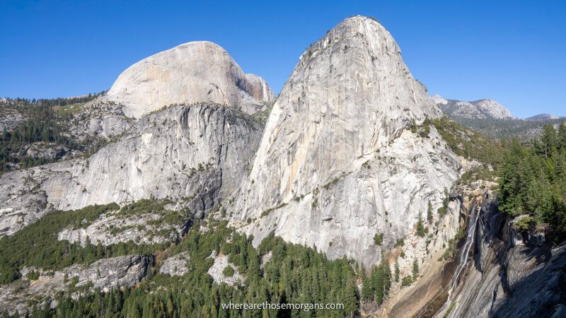 Photo of granite monoliths and domes bursting out of a rocky mountainous landscape filled with evergreen trees and a wispy waterfall in Yosemite