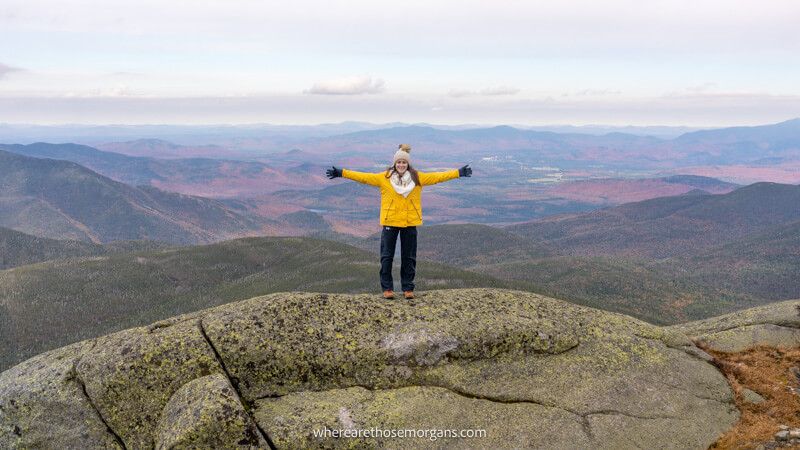 Photo of a hiker in a yellow coat stood on top of a rocky mountain summit with arms stretched out and wide open views behind