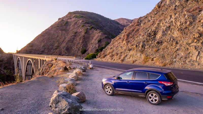 Photo of a car parked on a coastline near a bridge next to a headland at sunset