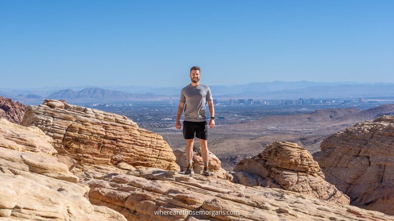 Photo of a hiker in shorts and t-shirt standing on a red rocky summit with views over Las Vegas in the distant background