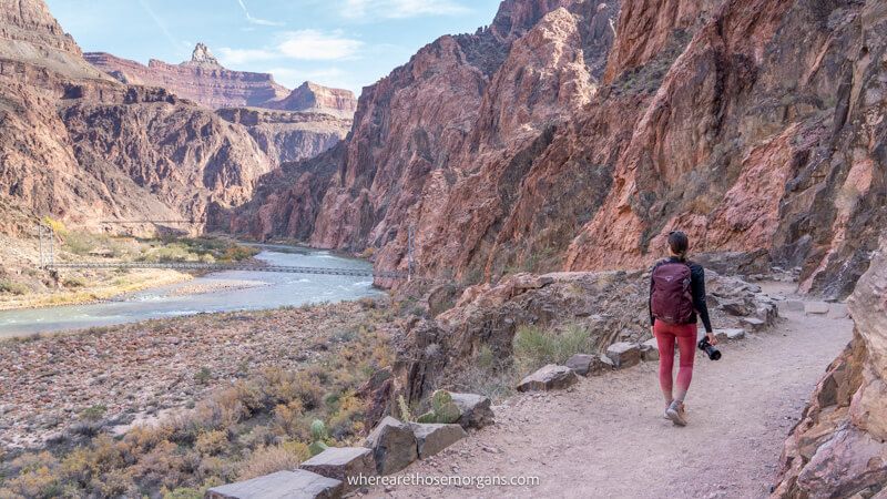 Photo of a person walking with a camera along a path next to a river and red rock cliff towards a metal bridge