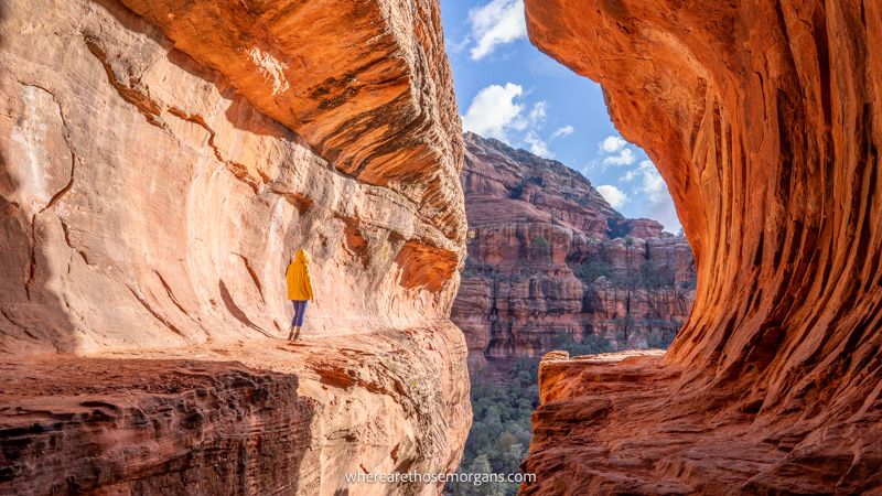 Photo of a hiker in yellow coat stood inside a subway tunnel shaped sandstone cave on a sunny day
