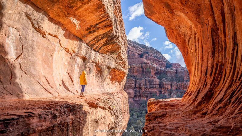 Photo of a person in a yellow coat stood inside a subway tunnel shaped sandstone cave looking out at views on a sunny day in Sedona AZ