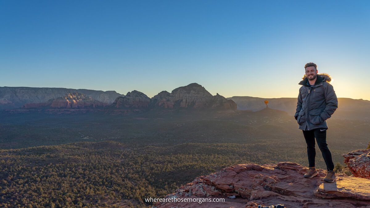 Tourist in thick winter coat standing on the edge of a red rock mesa with views over a valley behind at sunrise