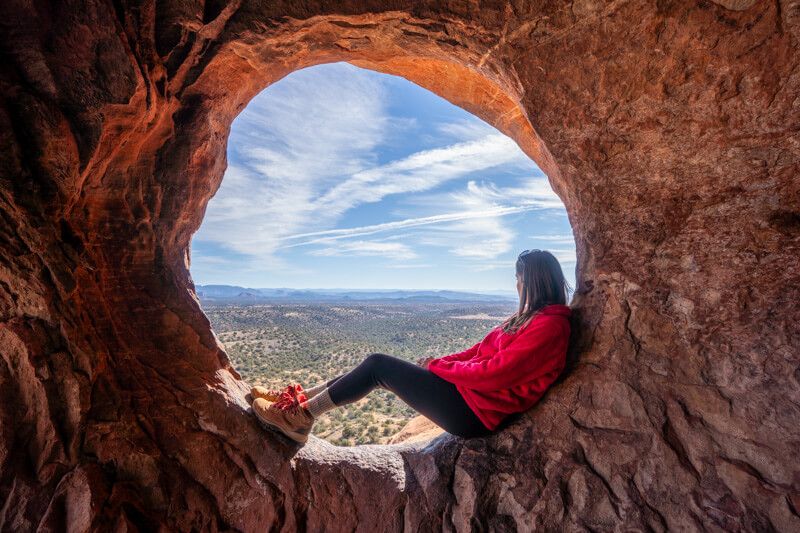 Photo of Kristen Morgan laying down inside a circular-shaped hole of a red rock cave wall looking at views over Sedona