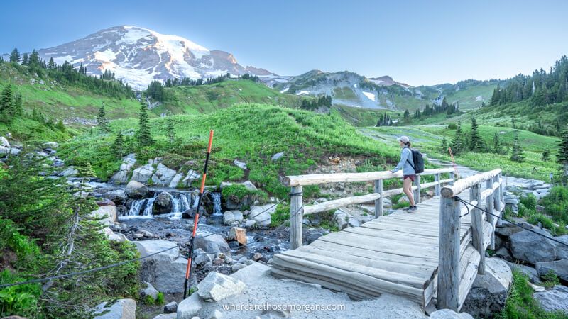 Photo of a person hiking over a wooden bridge to green meadows with a snow capped volcano behind at dawn