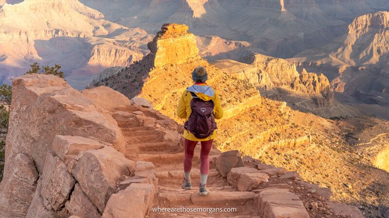 Photo of a hiker in pink pants and yellow coat walking down a stone staircase into the Grand Canyon at sunrise