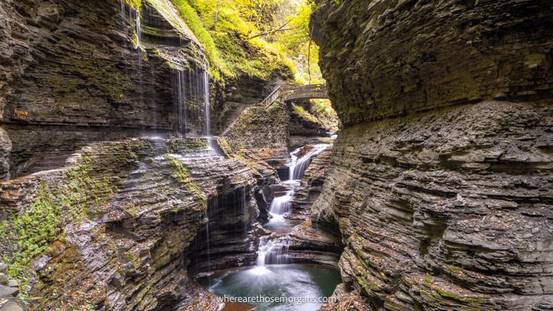 Photo of a rugged narrow gorge with a wispy waterfall flowing through and trees behind a stone bridge