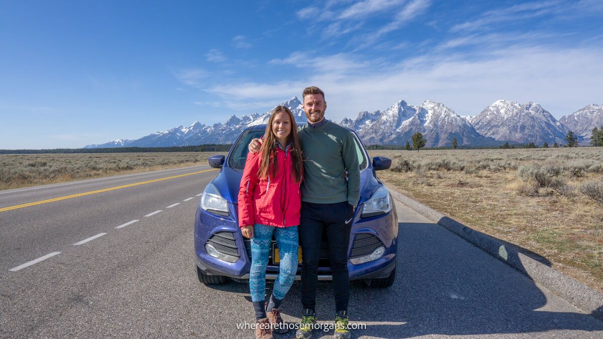 Photo of two tourists leaning against the front of a car in a wide open landscape with mountains in the background