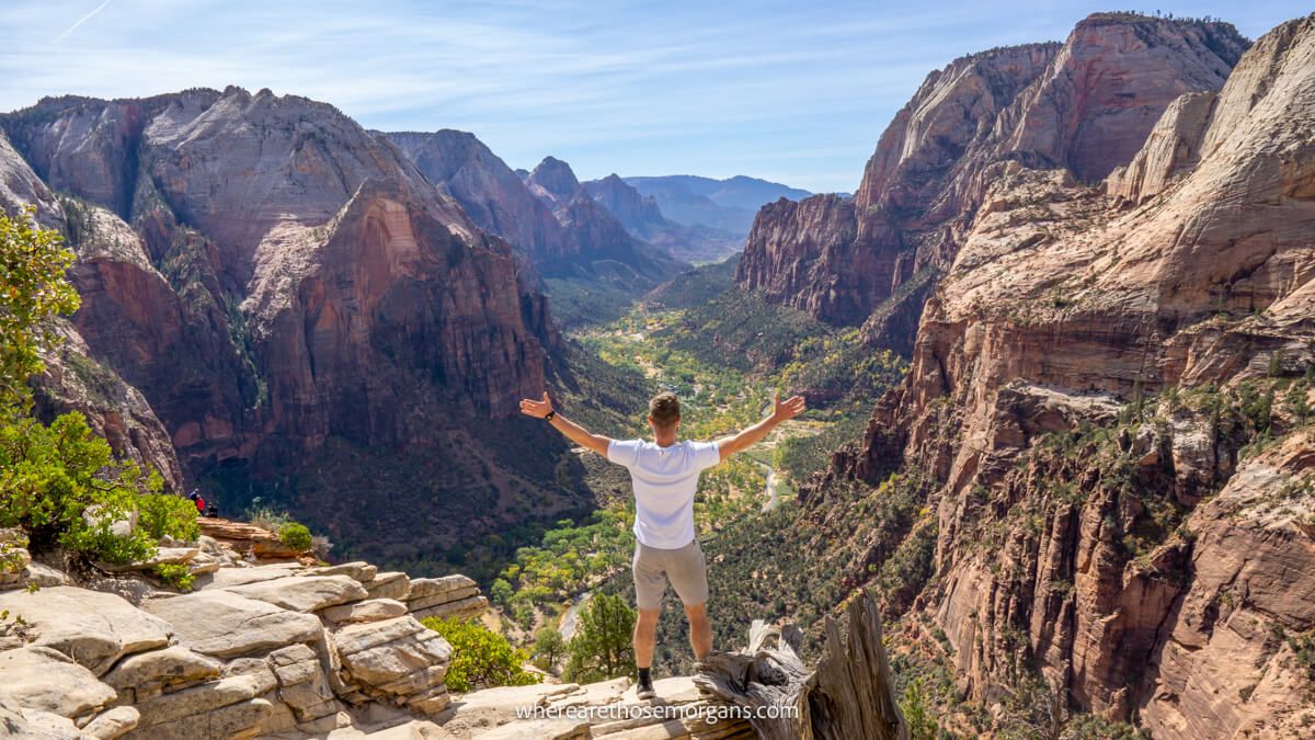 Photo of a tourist in white t-shirt with arms stretched out standing on a small rocky ledge looking out at views over a deep canyon with tall sandstone walls to either side