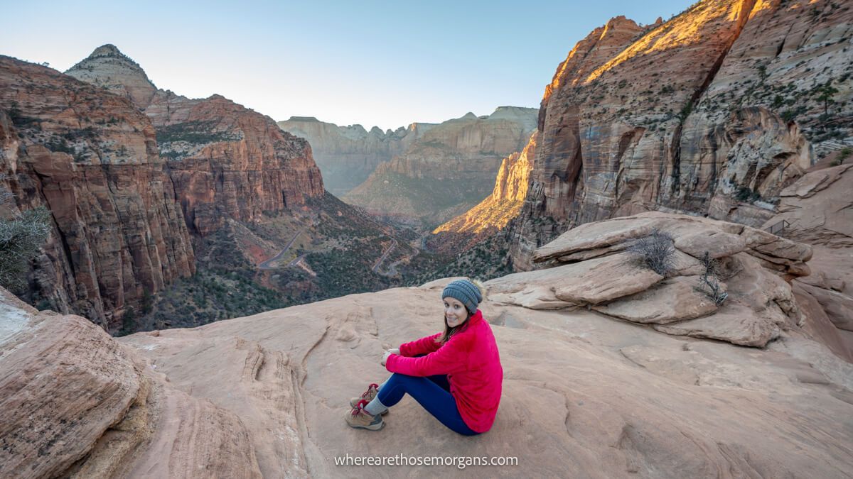 Photo of a tourist in pink fleece and hat sat on a rocky ledge in front of long drops into a deep canyon with orange sunlight glowing on tall red rock walls at sunset