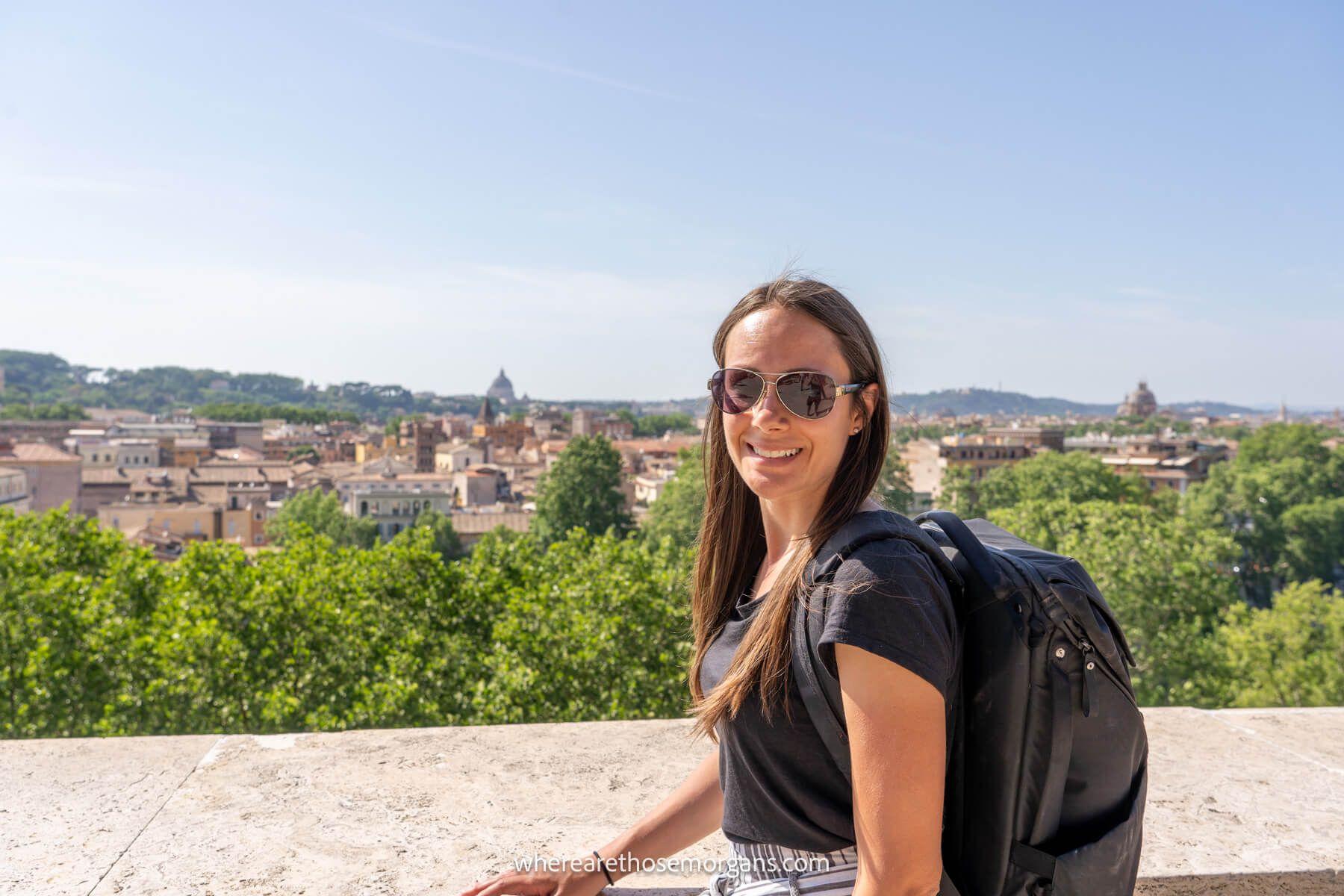 Photo of Kristen Morgan from Where Are Those Morgans standing on a terrace in front of a waist high wall with far reaching views behind of trees and Roman buildings on a clear day