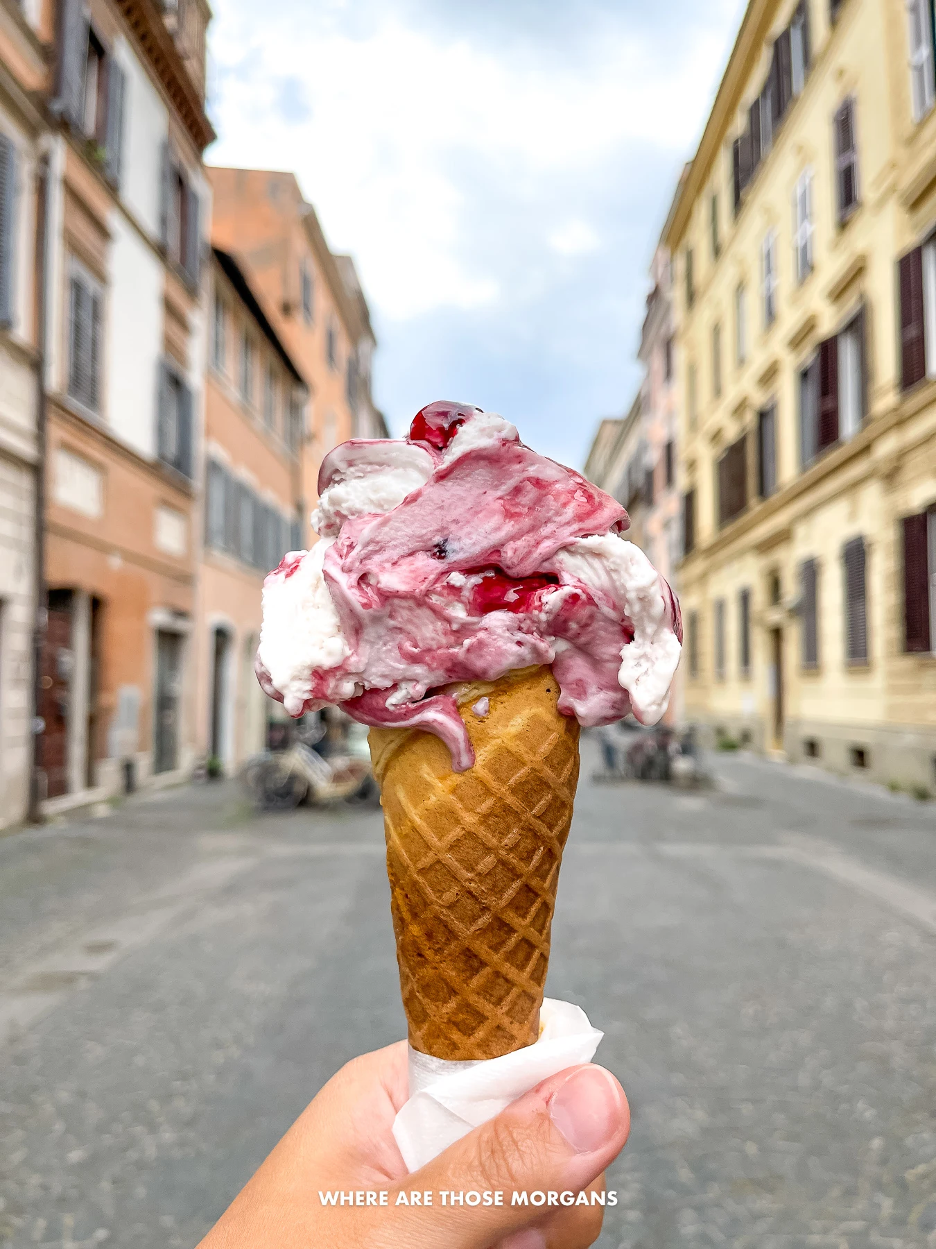 Hand holding an ice cream cone with pink and white ice cream in a narrow street with buildings either side