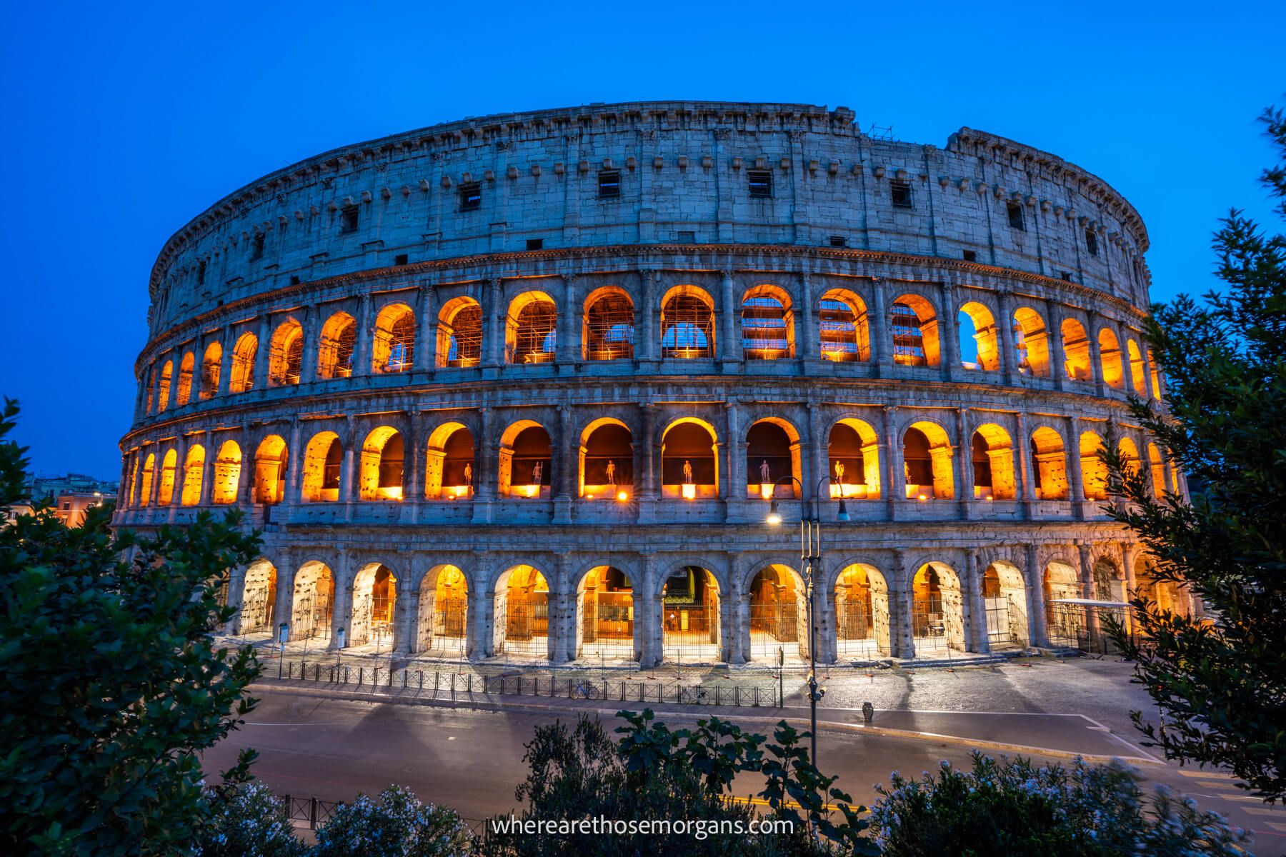 Photo of the Colosseum from a viewing area outside at night with trees in the foreground and a deep blue sky behind the orange lit up windows