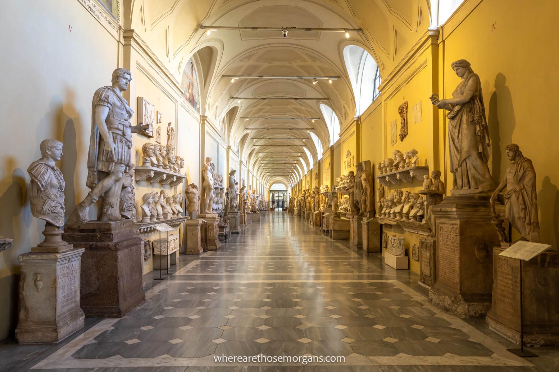 Photo of a hall inside a museum with no people inside and lots of sculptures on both sides of the hall, with yellow light illuminating the length of the corridor