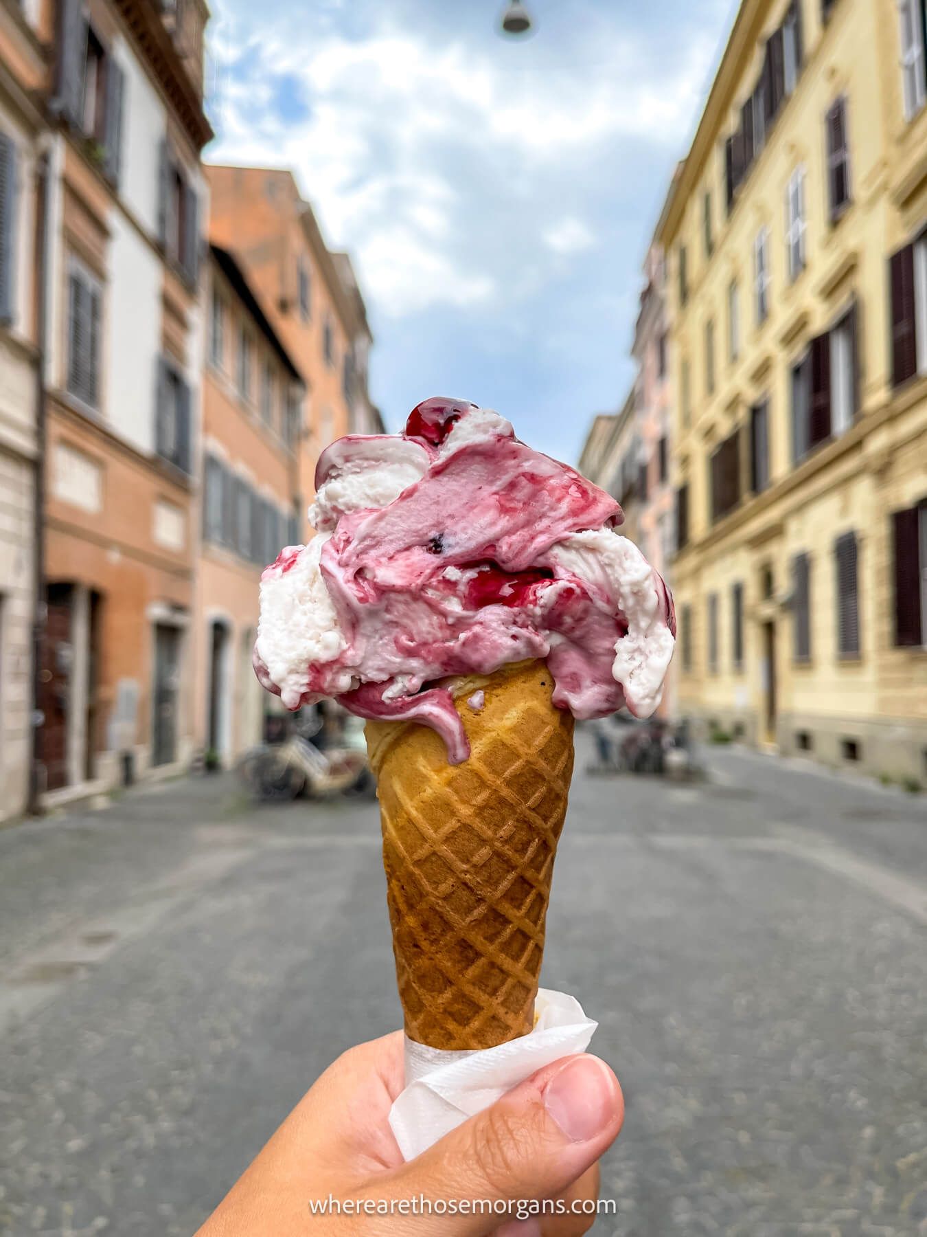 Photo of a pink and white gelato on top of a cone with a hand holding the ice cream in the middle of a narrow street with buildings