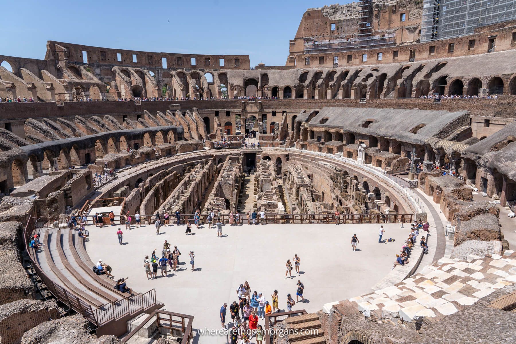 Photo of the inside of the Roman Colosseum with people walking on the arena under a clear blue sky