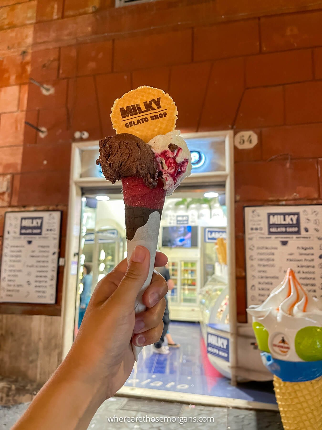 Photo of an arm and hand holding a gelato with biscuit saying milky at night outside a gelato shop