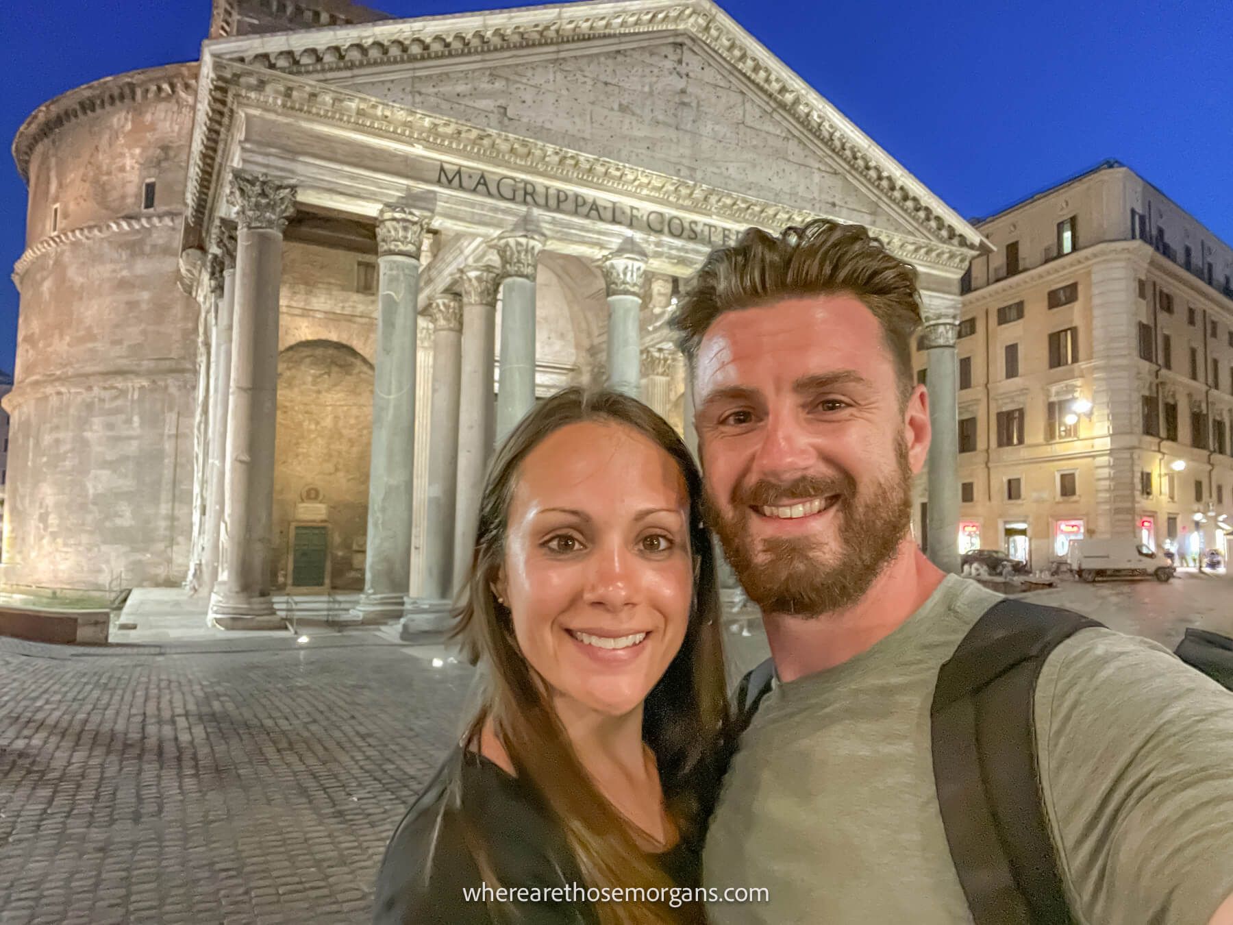 Photo of Mark and Kristen Morgan from Where Are Those Morgans taking a selfie in front of the Pantheon in Rome lit up at night against a deep blue sky