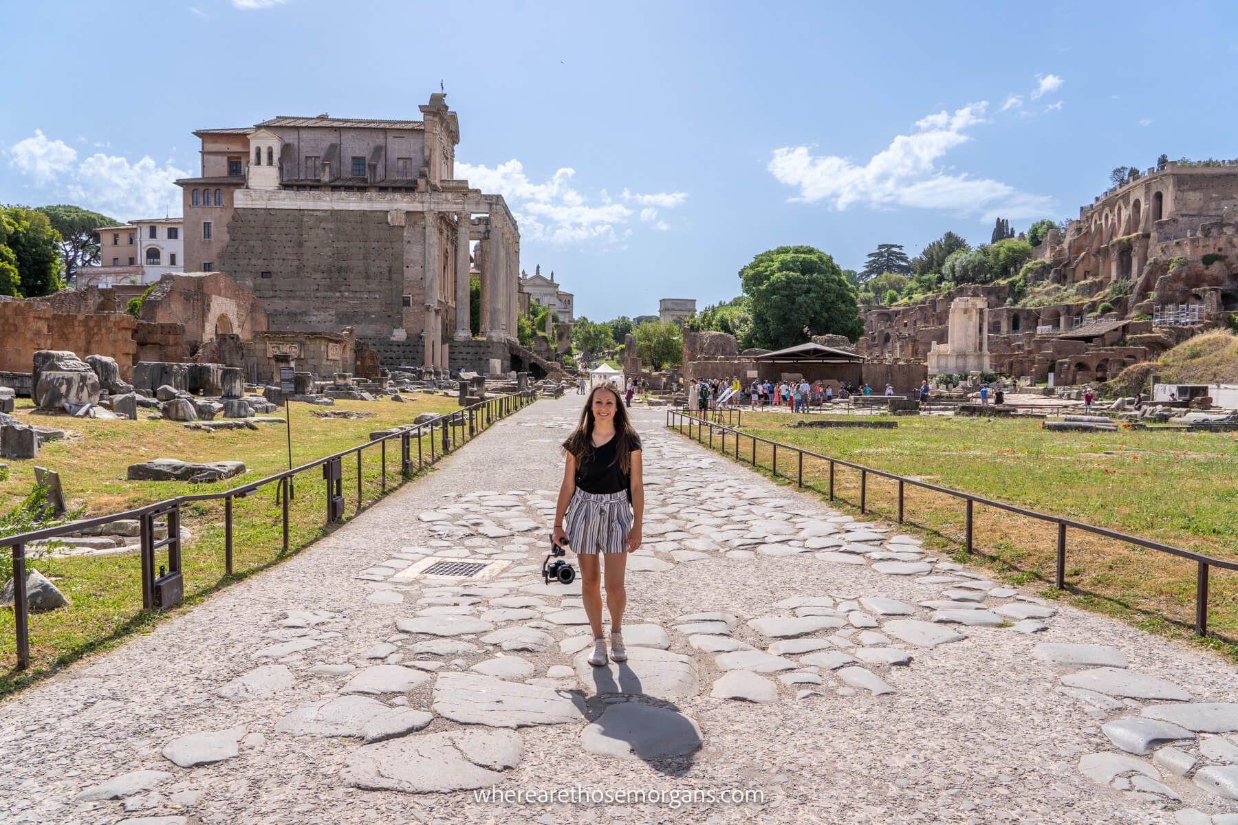 Photo of a tourist holding a camera stood on a stone path with grass to either side containing ancient ruins under a clear sky