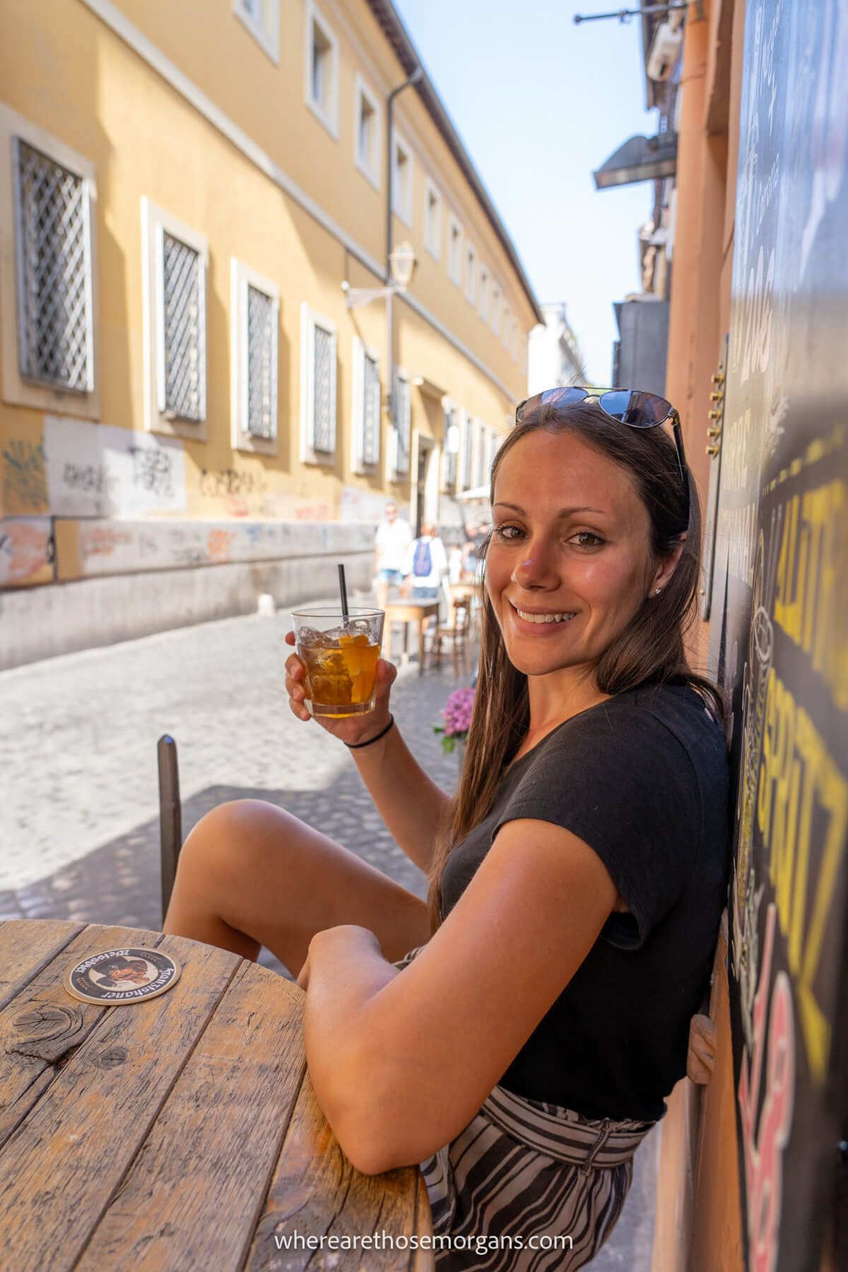 Photo of Kristen Morgan from Where Are Those Morgans sat at a small wooden table with her back to a wall in a narrow alley drinking a cocktail in Trastevere