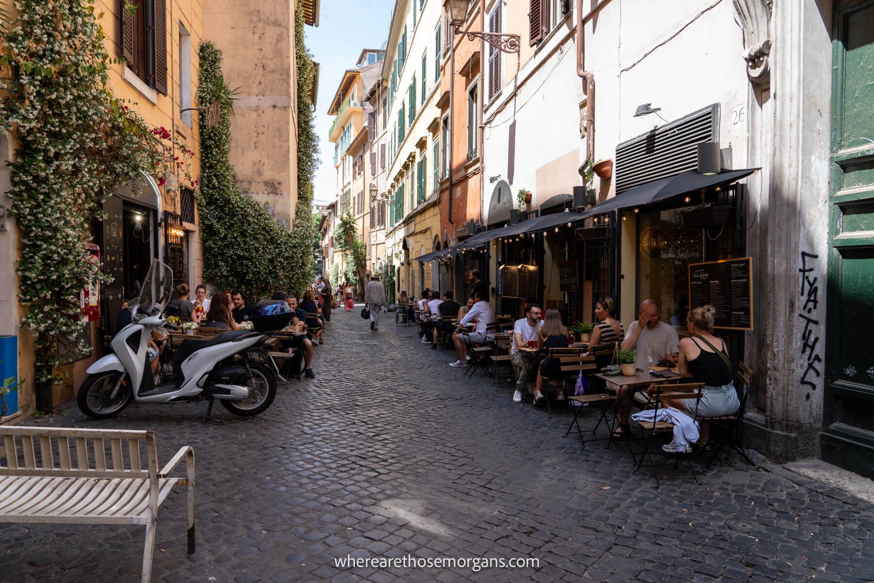 Photo of a narrow cobbled street with restaurants and tourists eating outside on a sunny day in Trastevere