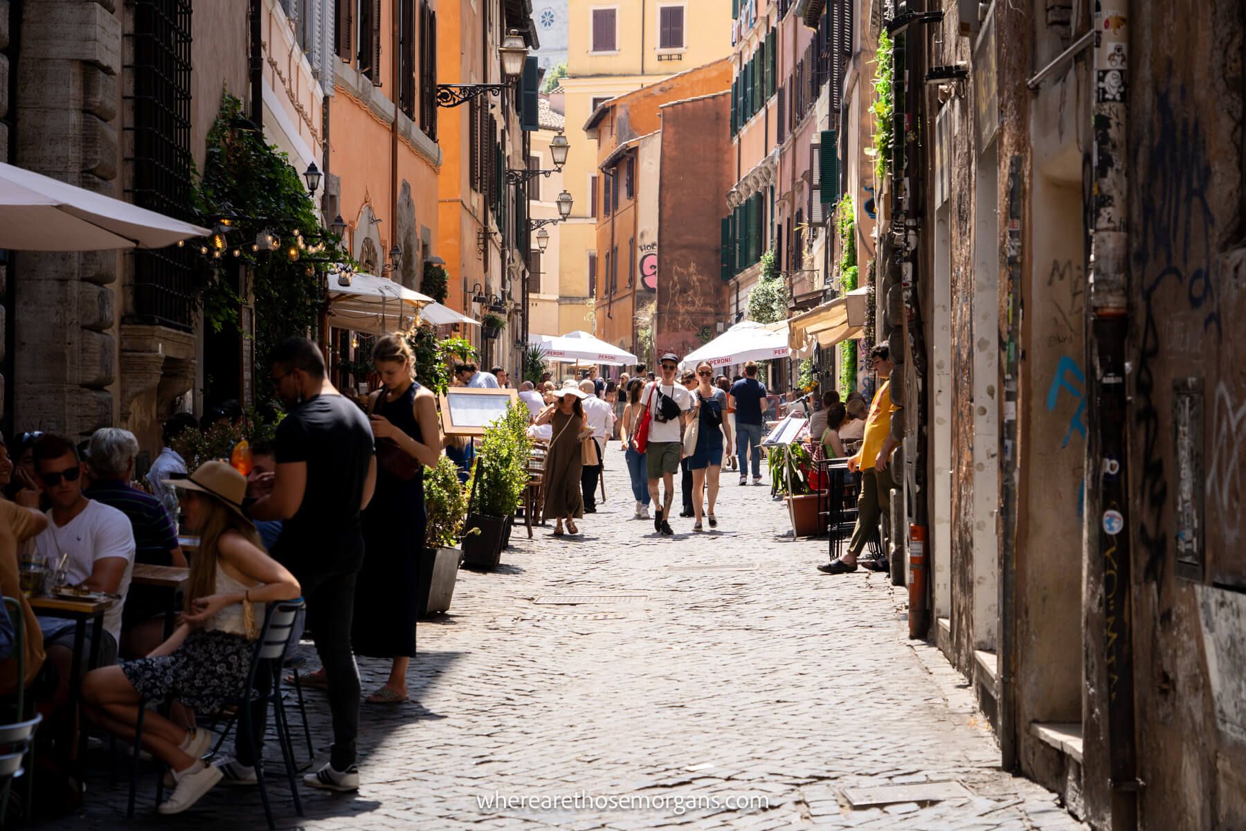 Photo of a narrow cobbled pedestrian street with tourists walking and eating at restaurants with outside tables on a sunny day in Trastevere in Rome