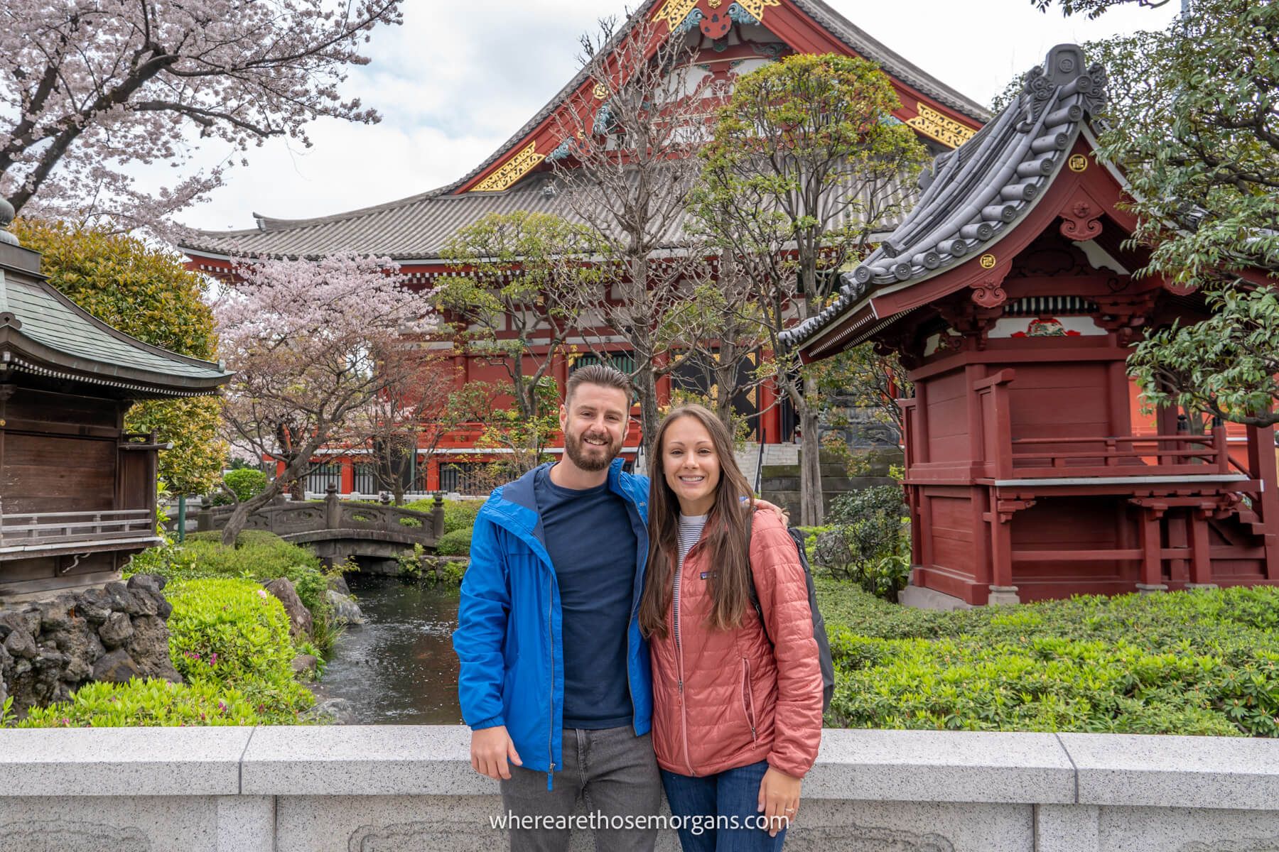 where are those morgans japan Photo of Mark and Kristen Morgan from Where Are Those Morgans standing together in front of a stone wall with green shrubs, cherry blossom trees and red wooden buildings at a temple in Japan
